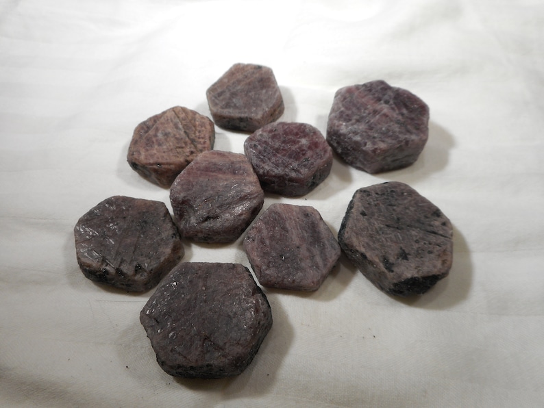 May include: A collection of nine faceted, dark purple gemstones. The gemstones are shaped like hexagons and have a rough, natural finish.