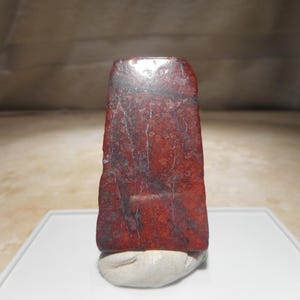 May include: A polished, rectangular stone slab with a vibrant red base color, featuring intricate dark grey and black veining patterns throughout the surface. The stone has a smooth, reflective finish and is supported by a small, light-colored base.