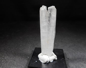 Pode incluir: Uma formação cristalina translúcida e clara, possivelmente selenite, está em pé sobre uma base preta. O cristal tem uma estrutura vertical e colunar com um topo dividido, mostrando texturas naturais e sutis variações de tom. A base é uma plataforma retangular escura.