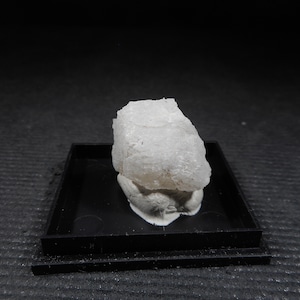 Peut inclure: Un spécimen minéral blanc et texturé est posé sur un plateau noir sur fond sombre. Le minéral a un aspect rugueux et cristallin, et le plateau offre une base contrastée pour l'objet.