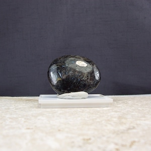 May include: A smooth, dark gray and black gemstone with a slight sheen. The gemstone is round and sits on a white pedestal.