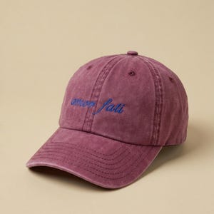 Könnte beinhalten: Burgunderfarbene Baseballkappe mit gebogenem Schirm. Auf der Kappe ist der Schriftzug "amor fati" in blauer Schreibschrift aufgestickt. Der Hut ist aus einem weichen, strukturierten Stoff gefertigt.