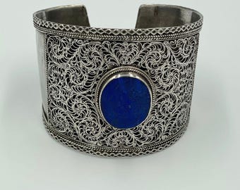 May include: Silver cuff bracelet with intricate filigree design and a large, round, deep blue stone in the center. The bracelet has a wide band with detailed patterns and a polished finish, suitable for a bold statement.
