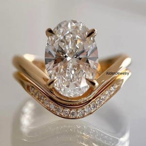 May include: A gold ring set featuring a large oval-cut center stone held by four prongs, paired with a curved band encrusted with small round diamonds. The metal has a polished finish, and the text AldenJewelry is visible on the side of the band.