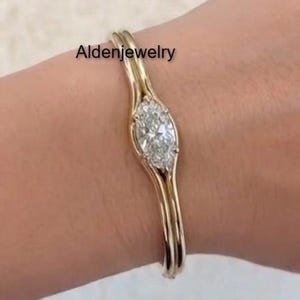 May include: A gold-colored bracelet with a central oval-shaped diamond. The bracelet has a delicate, curved design, with the diamond set in a prong setting. The text "Aldenjewelry" is visible on the image.