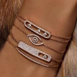 Könnte beinhalten: Armbänder aus Roségold mit funkelnden Diamantakzenten. Die Armbänder weisen verschiedene Designs auf, darunter eine rechteckige Form, eine Augenform und eine Stäbchenform, alle mit kleinen, glitzernden Diamanten verziert. Die Armbänder sind für einen stilvollen Look geschichtet.