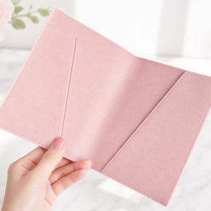 May include: A light pink passport holder featuring a textured finish and two diagonal interior pockets for document storage. The open cover is constructed from a durable material with clean stitched edges, providing a simple and functional design.