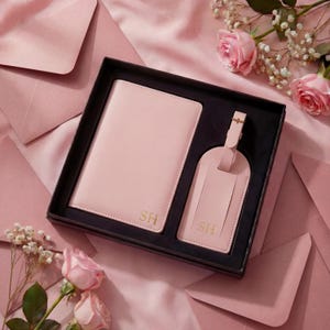May include: A light pink passport holder and a matching luggage tag set presented in a black gift box. Both items feature the gold embossed initials SH. The set is arranged on a soft pink fabric surface surrounded by light pink roses and envelopes.