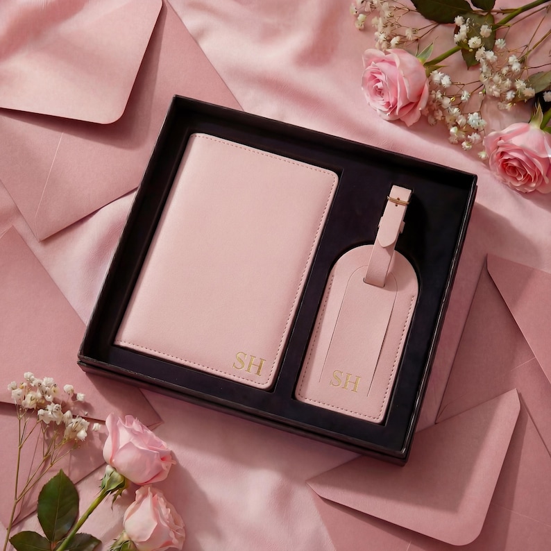 May include: A light pink passport holder and a matching luggage tag set presented in a black gift box. Both items feature the gold embossed initials SH. The set is arranged on a soft pink fabric surface surrounded by light pink roses and envelopes.