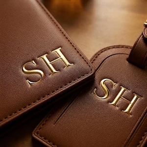 May include: A brown textured passport holder and a matching luggage tag, both featuring the gold embossed letters SH. The items have a cross-hatched surface finish and are positioned side by side, displaying the monogram clearly on each piece.
