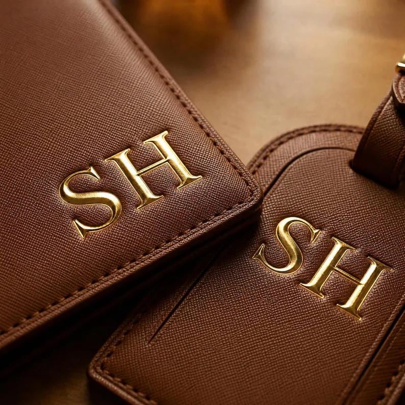 May include: A brown textured passport holder and a matching luggage tag, both featuring the gold embossed letters SH. The items have a cross-hatched surface finish and are positioned side by side, displaying the monogram clearly on each piece.