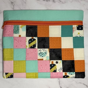 May include: A rectangular zippered pouch featuring a patchwork design of squares in pink, teal, black, orange, and cream. The fabric displays patterns of poison bottles, spiders, and dots. A solid teal fabric band runs across the top with an orange zipper.