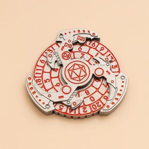 May include: A silver and red D&D dice spinner. The spinner has a circular design with numbers and dice notations like D20, D10, and D8. The center features a red D&D symbol. The design is intricate and detailed, with a metallic finish.