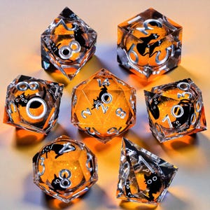 Peut inclure: Ensemble de sept dés polyédriques avec un cœur orange et un extérieur en résine transparente. Chaque dé présente des chiffres argentés et une silhouette de chat noir. Ces dés sont conçus pour les jeux de société et ont un thème Halloween.