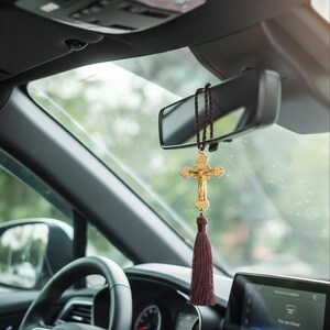 May include: A gold-colored crucifix car charm hanging from the rearview mirror. The crucifix features a detailed depiction of Jesus Christ and is adorned with a burgundy tassel. The car's interior includes a steering wheel and dashboard.