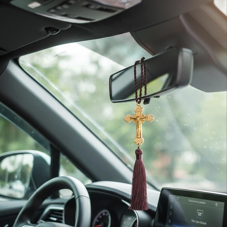 May include: A gold-colored crucifix car charm hanging from the rearview mirror. The crucifix features a detailed depiction of Jesus Christ and is adorned with a burgundy tassel. The car's interior includes a steering wheel and dashboard.