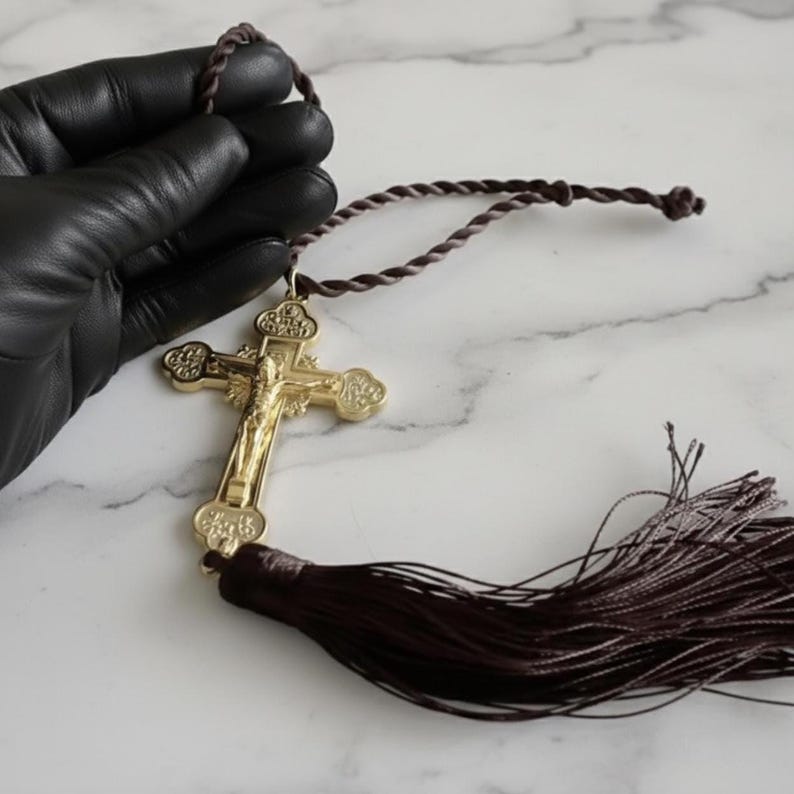 May include: A gold-colored crucifix with a detailed depiction of Jesus Christ, suspended from a twisted brown cord. A matching brown tassel hangs below the cross. The cross is held by a gloved hand.