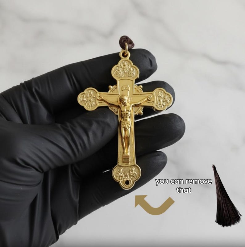 May include: A gold-colored crucifix with a detailed depiction of Jesus Christ. The cross has ornate detailing and is suspended from a brown cord. The image is set against a white marble background.