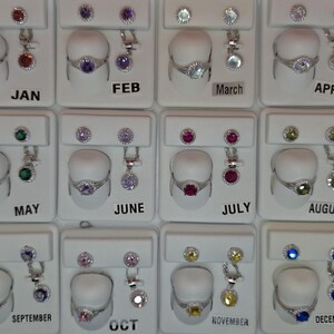May include: Jewellery display featuring rings, stud earrings, and pendant necklaces. Each set has a different coloured gemstone, with the months of the year labelled below each set. The gemstones include red, purple, blue, green, and yellow.
