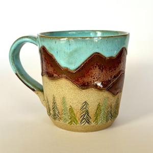 Puede incluir: Taza de cerámica hecha a mano con un diseño de montaña y bosque. La taza presenta un cielo azul turquesa, montañas marrones y árboles verdes. El asa y el borde también son de color azul turquesa. Una pieza única y rústica.