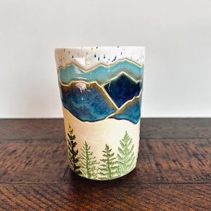 May include: A ceramic tumbler featuring a hand-painted mountain landscape with shades of deep blue and turquoise glaze at the top, transitioning into a cream-coloured base decorated with a series of green pine trees around the lower exterior.