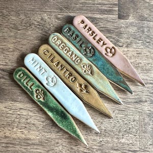 Puede incluir: Seis marcadores de jardín de cerámica en varios colores, incluyendo verde, rosa y beige. Cada marcador está etiquetado con el nombre de una hierba: Eneldo, Menta, Cilantro, Orégano, Albahaca y Perejil. Los marcadores tienen un extremo puntiagudo.