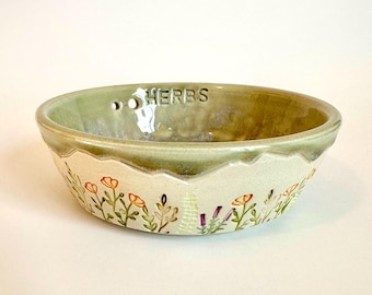 May include: A round ceramic bowl with a light green interior and a cream-colored exterior. The word HERBS is embossed on the inner rim. The exterior features hand-painted orange flowers, green foliage, and purple accents around the base.