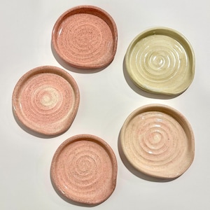 Può includere: Cinque piatti in ceramica fatti a mano, nei toni del rosa e del crema, sono disposti su una superficie bianca. Ogni piatto ha una forma circolare con un bordo leggermente rialzato e un motivo a spirale strutturato al centro. I piatti variano leggermente per colore e dimensione.