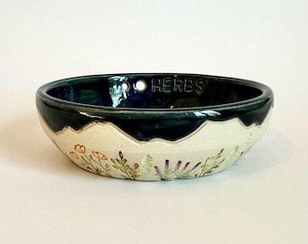 May include: A shallow ceramic bowl with a dark blue glazed interior and a cream-colored exterior. The exterior features hand-painted botanical illustrations of orange and purple flowers. The word HERBS is embossed on the inner rim.