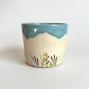 May include: A ceramic cup featuring a smooth, glossy turquoise glaze on the upper portion and a matte, off-white base. The lower section is decorated with hand-painted botanical illustrations of green foliage, purple sprigs, and orange flowers.