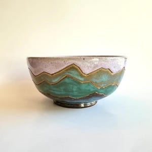 Può includere: Una ciotola in ceramica con un design a forma di montagna. La ciotola presenta una parte superiore color lavanda, una sezione centrale turchese e una base blu scuro. Il design della montagna è delineato in oro. È un pezzo decorativo.