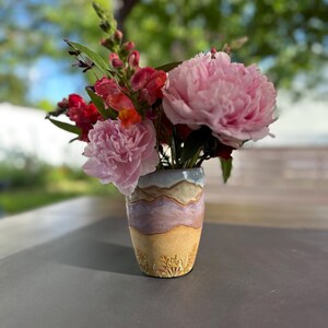May include: A ceramic vase with a unique design holds a vibrant bouquet of flowers. The vase features a textured, layered design in shades of tan, lavender, and blue. The bouquet includes pink peonies and red snapdragons, creating a colorful floral arrangement.