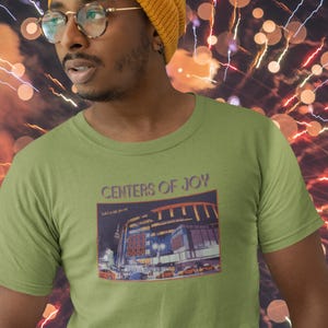 May include: A light green t-shirt with the text "CENTERS OF JOY" and a graphic of a building at night. The person in the image is wearing a yellow beanie and glasses. The background features bokeh lights and fireworks.