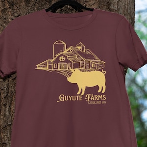 Puede incluir: Una camiseta granate con una ilustración dorada de una granja, un cerdo y el texto "GUYUTE FARMS ESTABLISHED 1994". La camiseta está colgada de una percha negra contra un tronco de árbol.