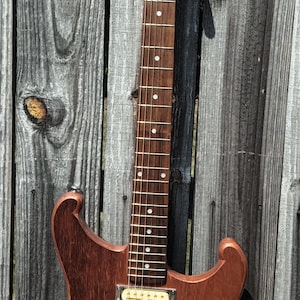 Puede incluir: Una guitarra eléctrica de madera con acabado natural, con dos pastillas, herrajes cromados y una pala con las letras "BGL". La guitarra está apoyada contra una valla de madera desgastada.