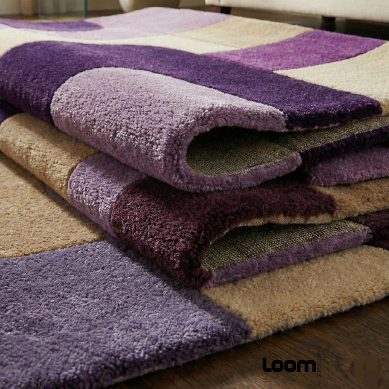 Puede incluir: Una alfombra mullida con un patr&oacute;n geom&eacute;trico en tonos morados y beige. La alfombra est&aacute; hecha de un material suave y texturizado y tiene una secci&oacute;n enrollada, revelando el reverso. El logotipo de Loom es visible en la esquina inferior derecha.