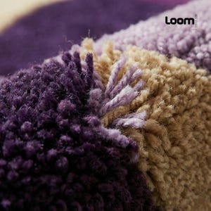Puede incluir: Primer plano de una alfombra texturizada con un dise&ntilde;o de bloques de color. La alfombra presenta secciones de morado oscuro, morado claro y beige. Las fibras son gruesas y afelpadas, creando una apariencia suave y acogedora. La palabra "Loom" es visible.