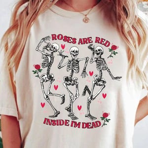 May include: Cream-colored t-shirt featuring a graphic of three dancing skeletons with red roses and hearts. The text "Roses are Red, Inside I'm Dead" is printed in red. The shirt has short sleeves.