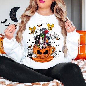 Op de afbeelding: Witte sweatshirt met een Halloween-thema. De afbeelding toont personages uit "The Nightmare Before Christmas" en Mickey Mouse-vormige ballonnen, die uit een oranje pompoen-theekop komen. Zwarte vleermuizen en spinnenwebben maken het ontwerp af.