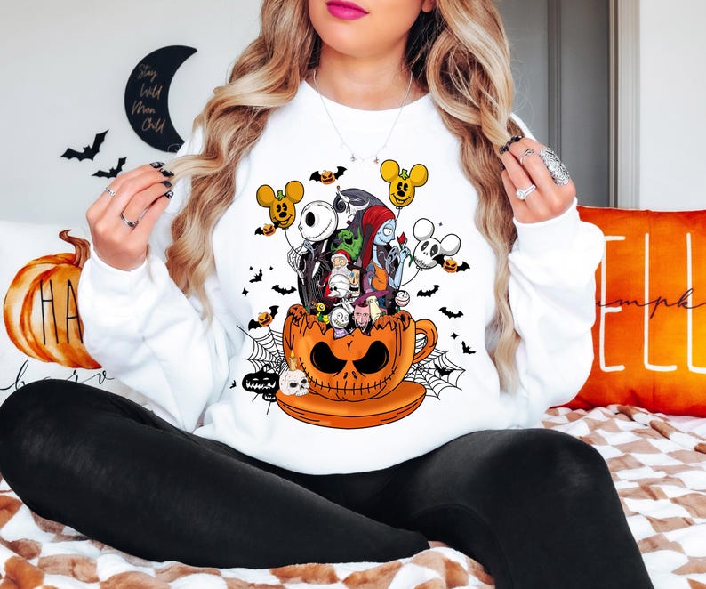 Op de afbeelding: Witte sweatshirt met een Halloween-thema. De afbeelding toont personages uit "The Nightmare Before Christmas" en Mickey Mouse-vormige ballonnen, die uit een oranje pompoen-theekop komen. Zwarte vleermuizen en spinnenwebben maken het ontwerp af.