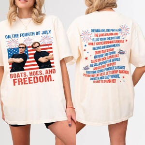 May include: Cream-colored t-shirt with a graphic on the front featuring two men in sunglasses in front of an American flag and the text "ON THE FOURTH OF JULY, BOATS. HOES. AND FREEDOM." The back of the shirt has a humorous poem.