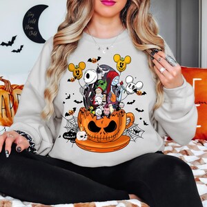 Op de afbeelding: Een lichtgrijs sweatshirt met een Halloween-thema. De afbeelding toont personages uit The Nightmare Before Christmas en Mickey Mouse, die uit een oranje pompoenbeker komen. Vleermuizen en spinnenwebben maken het spookachtige ontwerp compleet.