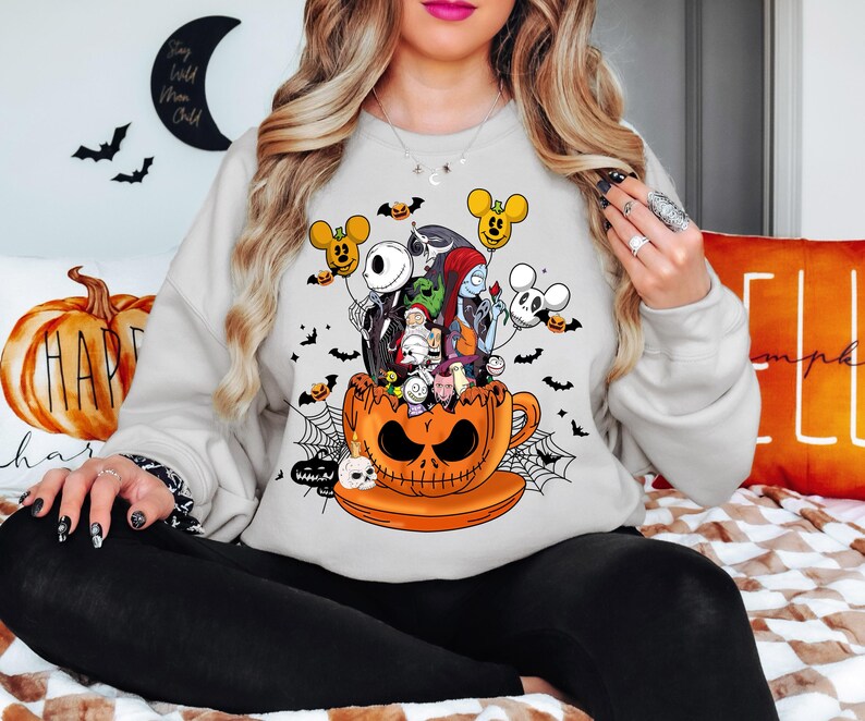 Op de afbeelding: Een lichtgrijs sweatshirt met een Halloween-thema. De afbeelding toont personages uit The Nightmare Before Christmas en Mickey Mouse, die uit een oranje pompoenbeker komen. Vleermuizen en spinnenwebben maken het spookachtige ontwerp compleet.