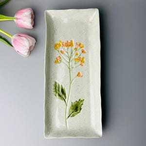 May include: A rectangular, off-white ceramic serving dish with a raised floral design in yellow and orange. The dish has a speckled texture and irregular edges. Green leaves and stem are also part of the floral design. Pink tulips are in the background.
