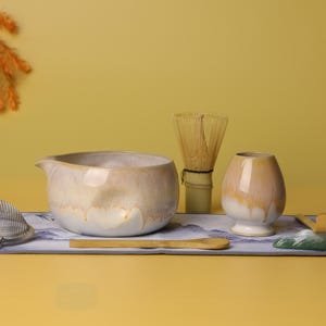 May include: A matcha tea set featuring a ceramic bowl, whisk, and tea cup. The set includes a metal strainer, a bamboo scoop, and a decorative mountain-shaped piece. The items are arranged on a patterned mat.