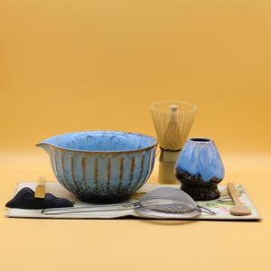 May include: A matcha tea set with a blue ceramic bowl, a bamboo whisk, a ceramic vase, a tea strainer, and a wooden spoon. The set is arranged on a decorative tray against a yellow background.