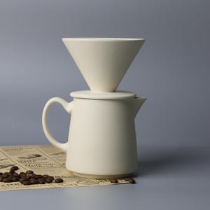 May include: A cream-coloured ceramic pour-over coffee maker, featuring a cone-shaped filter atop a small jug. The coffee maker rests on a newspaper-style sheet, with coffee beans scattered nearby. The background is a solid grey.