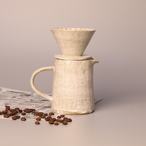 Op de afbeelding: Een crèmekleurige keramische pour-over koffiemaker met een getextureerd oppervlak. De set bevat een karaf, een deksel en een kegelvormig filter. Koffiebonen liggen verspreid in de buurt. De koffiemaker staat op een krant.