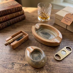 May include: A collection of brown ceramic ashtrays in various shapes, a cigar, a gold cigar cutter, a glass of whiskey, and a gift box with a "Happy Father's Day" tag. The items are arranged on a wooden surface.