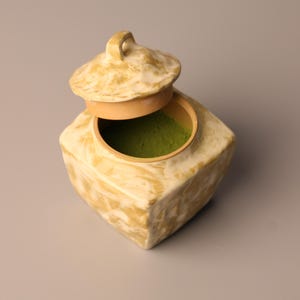 May include: A square, ceramic container with a lid, partially open to reveal a vibrant green powder inside. The container has a light beige, marbled finish. The lid has a small handle. The container is likely for storing tea or spices.